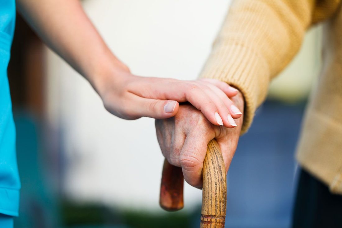 A carer steadying an older person's hand as they walk with a stick